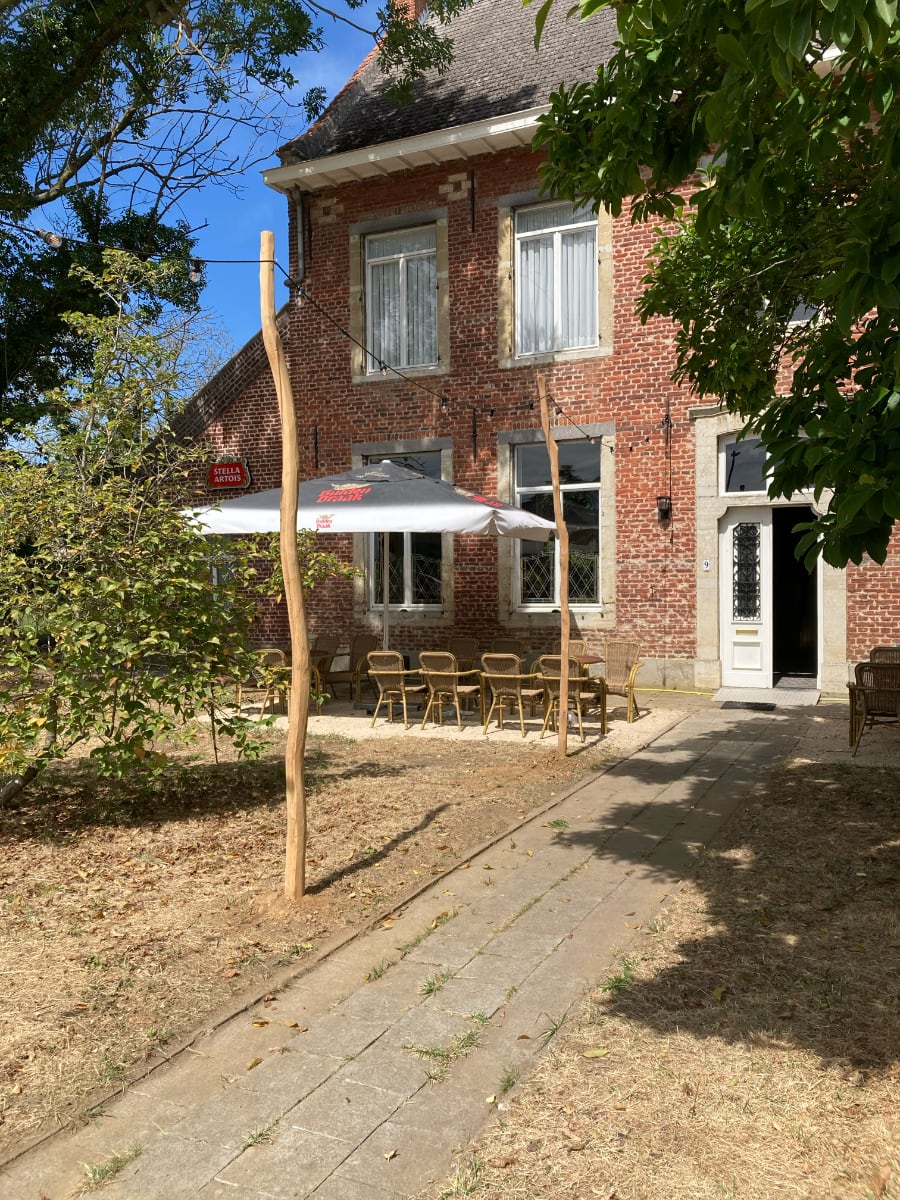 De voorkant van het Pastorie-gebouw, met een terras met tafels en een grote parasol op een zonnige dag.