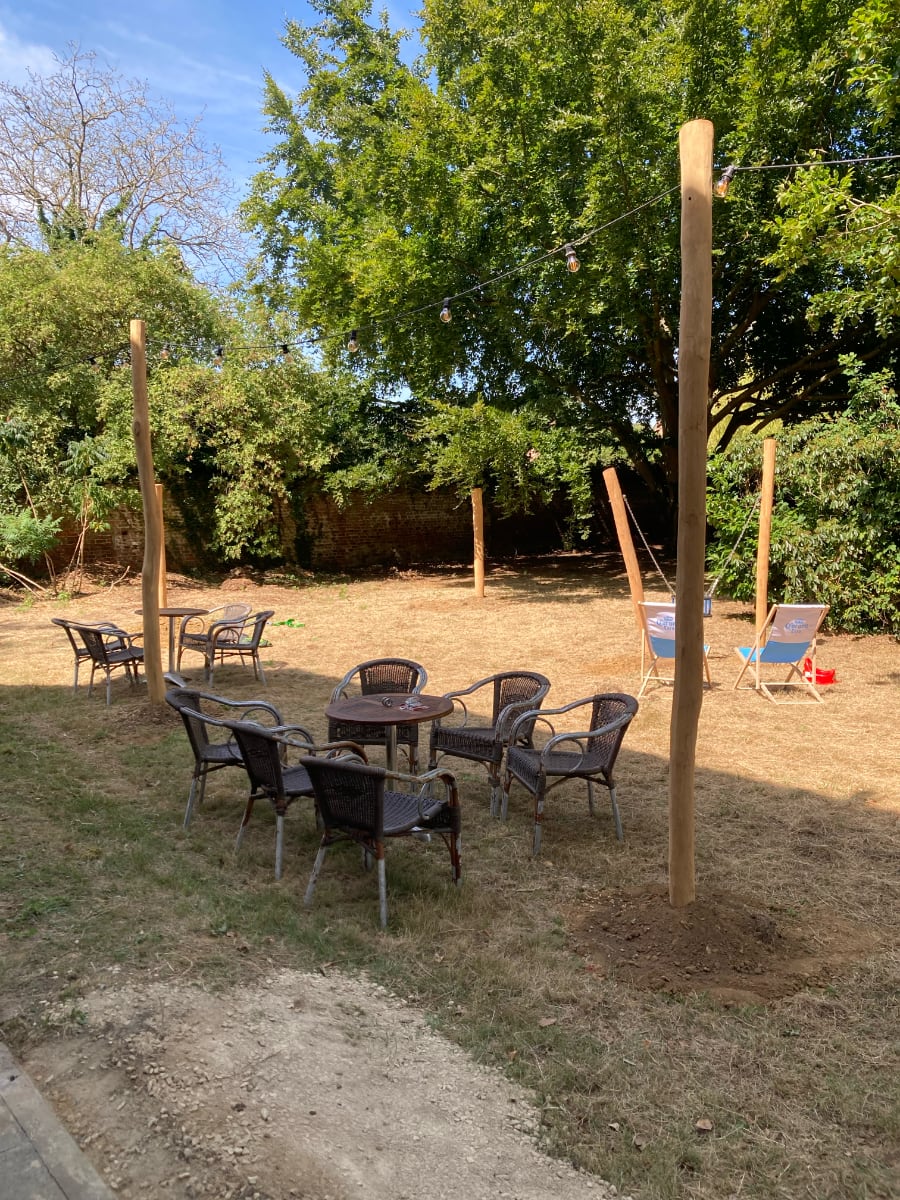 Een zonnige tuin van de Pastorie met meerdere zithoeken en stoelen, omringd door bomen en een bakstenen muur, met verlichting die tussen houten palen hangt.