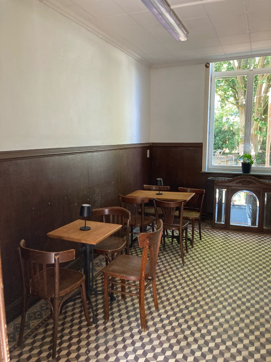 Een interieur van de Pastorie met twee houten tafels en stoelen, donkere houten lambrisering en een tegelvloer met een ruitpatroon, met uitzicht op de tuin door een groot raam.