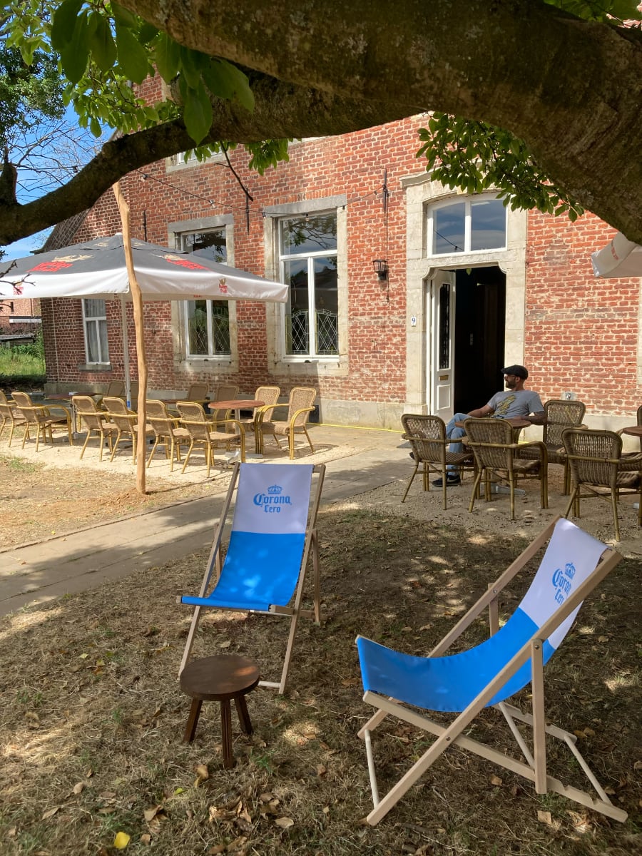 Het terras aan de voorgevel van de Pastorie, met rieten tafels en stoelen, een grote parasol en twee blauwe strandstoelen.