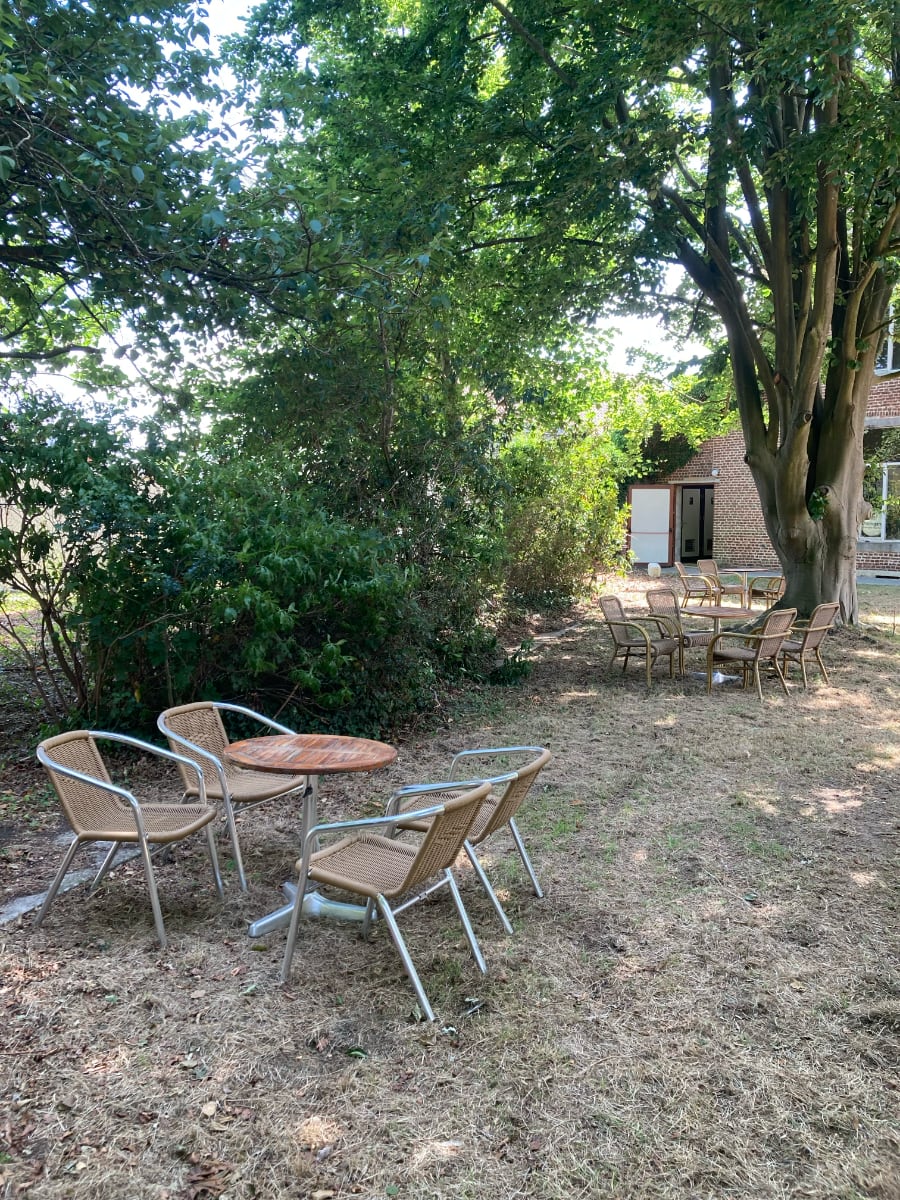Twee zithoeken in de achtertuin van de Pastorie, elk met een ronde tafel en rieten stoelen, geplaatst op een grasveld omringd door bomen en struiken.